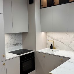 KoNad Group sp. z o.o. - Jasna kuchnia z marmurowym blatem i złotymi akcentami: bateria, uchwyty. Górne szafki z podświetleniem LED. Nowoczesny design z płytą indukcyjną i piekarnikiem.