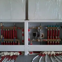 Rozdzielacz ogrzewania podłogowego w białej obudowie, z widocznymi rurami w izolacji termicznej i pianką montażową, ułożony na szarej płycie.