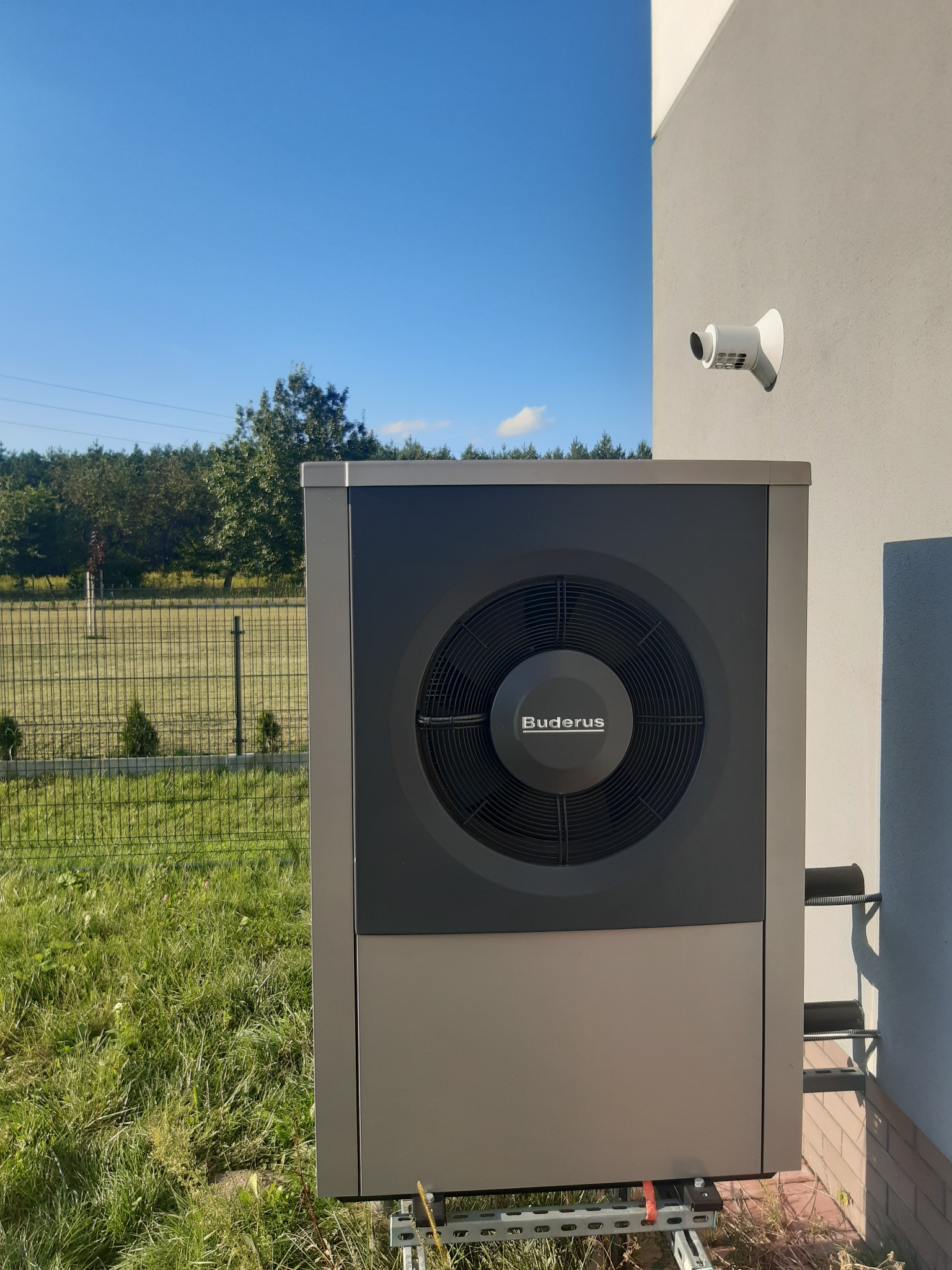 Szara pompa ciepła monoblok marki Buderus zamontowana na zewnątrz budynku, widoczny fragment elewacji, elementy mocujące i trawnik w słoneczny dzień.