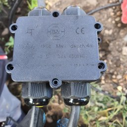 Elek-Info-Sys - Czarna puszka elektryczna IP68 z certyfikatami TUV i CE, podłączona do dwóch kabli, trzymana w dłoni w rękawicy roboczej na tle ziemi i trawy.