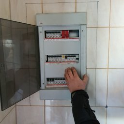 Elek-Info-Sys - Otwarta skrzynka elektryczna ABB z bezpiecznikami na kafelkowanej ścianie. Widoczna dłoń osoby sprawdzającej instalację. Gniazdko z kablem w dolnym rogu.
