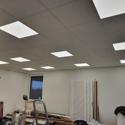 Elek-Info-Sys - Kwadratowe panele LED w suficie podwieszanym, widoczne drabiny i materiały budowlane w pomieszczeniu w trakcie prac wykończeniowych. Oświetlenie biurowe.