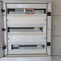 Elek-Info-Sys - Rozdzielnica elektryczna z bezpiecznikami i wyświetlaczem, zamontowana na kafelkowanej ścianie. Widoczna otwarta obudowa i elementy instalacji.