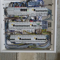 Elek-Info-Sys - Rozdzielnia elektryczna w białej obudowie, widoczne zabezpieczenia Eaton, przewody w peszlach i oznaczenia. Instalacja po montażu na szarym, surowym tle ściany.