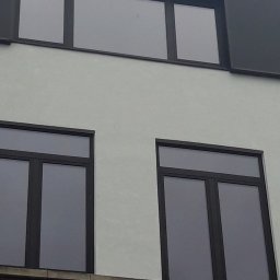 Fenetrex - Elewacja budynku z trzema oknami o ciemnych ramach. Dwa okna dwuskrzydłowe na dole, jedno trójdzielne u góry. Jasna ściana, ciemne obramowanie dachu.