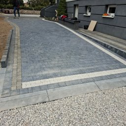 Ka.Myk-TRANS - Ułożona szara kostka brukowa z jasnymi pasami, obrzeża z kostki. Widok na podjazd do domu z szarym budynkiem w tle. Osoba w oddali. Żwir na obrzeżach.