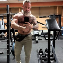 Maciej Chojnicki - Mężczyzna z brodą i tatuażami prezentuje muskulaturę, robiąc selfie w siłowni. Widoczne maszyny do ćwiczeń i ciężary. Ubrany w sportowe spodnie i buty.