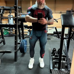 Maciej Chojnicki - Mężczyzna z brodą w sportowym stroju z motywem pająka, robiący selfie w siłowni. W tle sprzęt do ćwiczeń i ciężary.