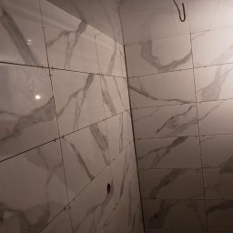 Armin Medynski - Ściana wyłożona białymi płytkami imitującymi marmur, z krzyżykami dystansowymi, w trakcie prac wykończeniowych w łazience. Widoczny fragment instalacji.