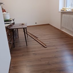 Armin Medynski - Pokój po położeniu paneli podłogowych. Widoczne listwy przypodłogowe, grzejnik i meble. Częściowo widoczne narzędzia i materiały budowlane.