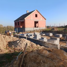 Armin Medynski - Budowa domu jednorodzinnego w stanie surowym otwartym: widoczne fundamenty z bloczków betonowych, ściany z czerwonej cegły i częściowo wykonany dach. Na pierwszym planie hałdy ziemi.