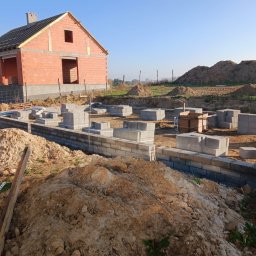 Armin Medynski - Budowa domu jednorodzinnego w Chełmcach: wzniesione fundamenty z bloczków betonowych, ściany z czerwonej cegły w stanie surowym otwartym, błękitne niebo.