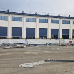 PROFDOOR - Nowoczesny budynek przemysłowy z segmentowymi bramami garażowymi w kolorze granatowym. Na pierwszym planie teren budowy z kałużami i sprzętem.
