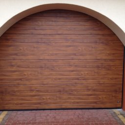 PROFDOOR - Zamknięta brama garażowa segmentowa w kolorze drewna, z łukowatym nadprożem, na tle kostki brukowej. Widoczny fragment elewacji budynku.