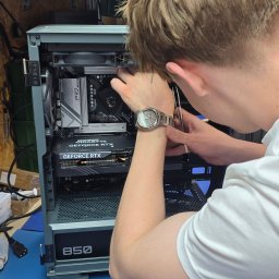 LunarTech Serwis Michał Bojarski - Serwis komputerowy Zielona Góra - Serwisant w zegarku montuje kartę graficzną GeForce RTX wewnątrz obudowy komputera PC z płytą główną ASRock Pro Series, zasilacz 850W, na niebieskim blacie.