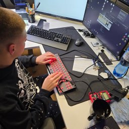LunarTech Serwis Michał Bojarski - Serwis komputerowy Zielona Góra - Serwisant testuje klawiaturę mechaniczną z podświetleniem LED na biurku z dwoma monitorami. Widoczne narzędzia diagnostyczne i notatnik.