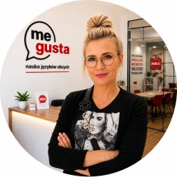 ME GUSTA NAUKA JĘZYKÓW OBCYCHCH ANNA MICHNO - Kobieta w okularach z upiętymi włosami stoi przed recepcją szkoły językowej 'me gusta', oferującej kursy języków obcych. W tle sala lekcyjna z czerwonymi krzesłami.