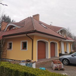 F. H. U. Bieniecki Kamil Bieniecki - Żółta elewacja domu z brązową dachówką i garażem z roletami, widoczne palety z dachówkami, samochód i fragment ogrodu w pochmurny dzień.