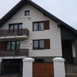 F. H. U. Bieniecki Kamil Bieniecki - Dwukondygnacyjny dom z jasną elewacją, ciemnymi elementami wykończeniowymi, drewnianymi balustradami balkonów i ogrodzeniem z białymi słupkami.