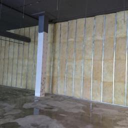 F. H. U. Bieniecki Kamil Bieniecki - Wnętrze w trakcie prac wykończeniowych: ściana z izolacją z wełny mineralnej zamontowana na metalowym stelażu, mokra betonowa podłoga, pracownik z mopem w tle.