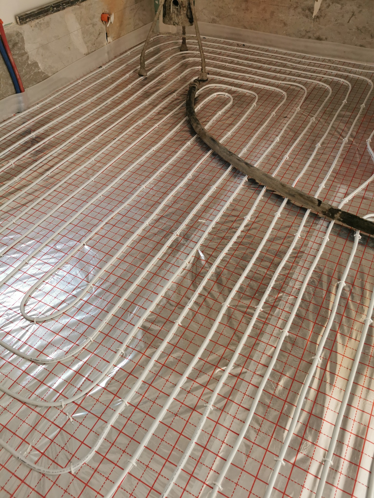 Rozłożone rury ogrzewania podłogowego na folii z czerwoną siatką, widoczny fragment ściany w stanie surowym i rura doprowadzająca zaprawę.
