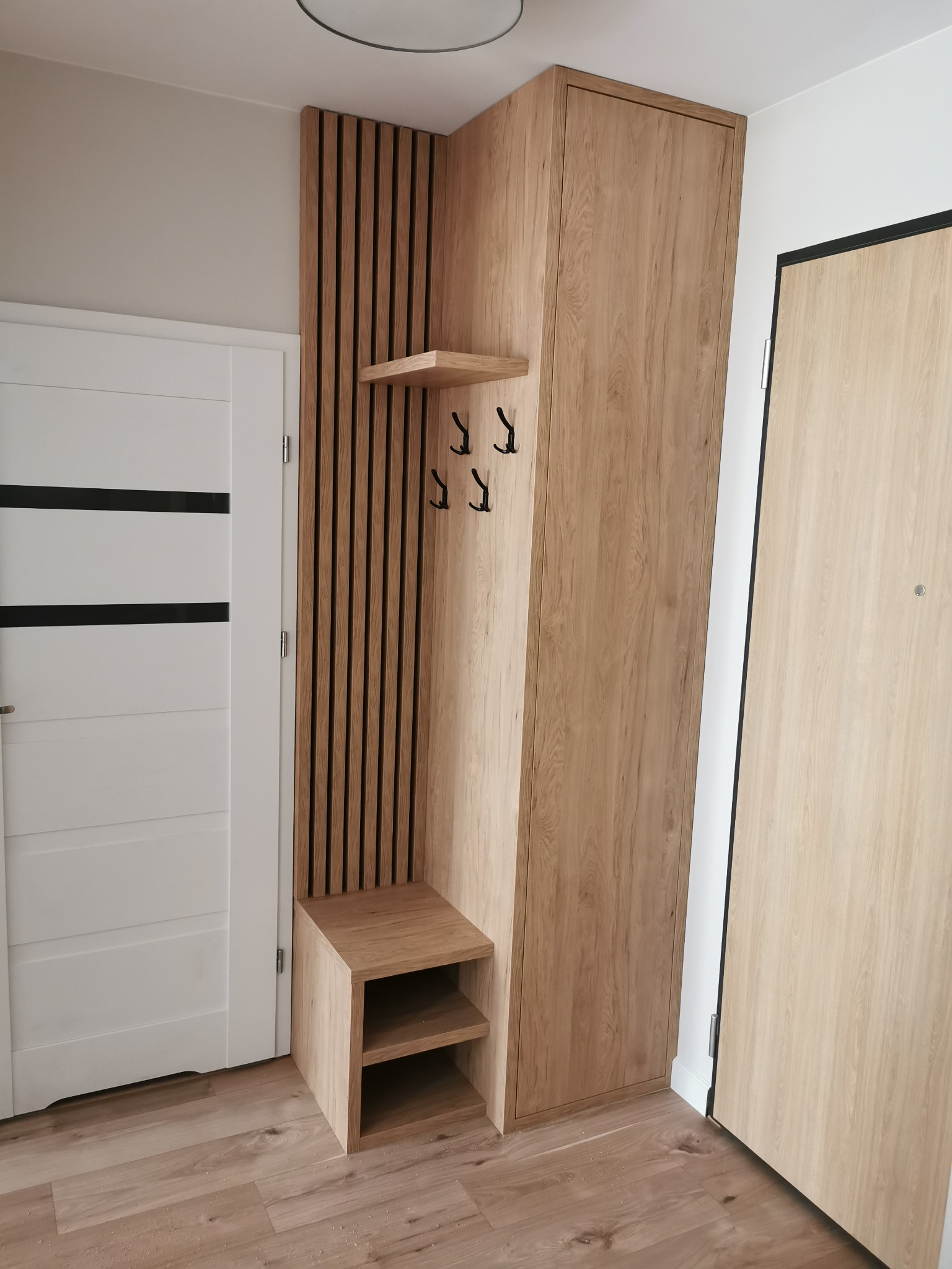 Wnętrze przedpokoju z zabudową meblową z drewna w odcieniach beżu, obejmującą szafę, panel z wieszakami i siedziskiem z półkami, sąsiadującą z białą ścianą i dwiema parami drzwi w podobnej...