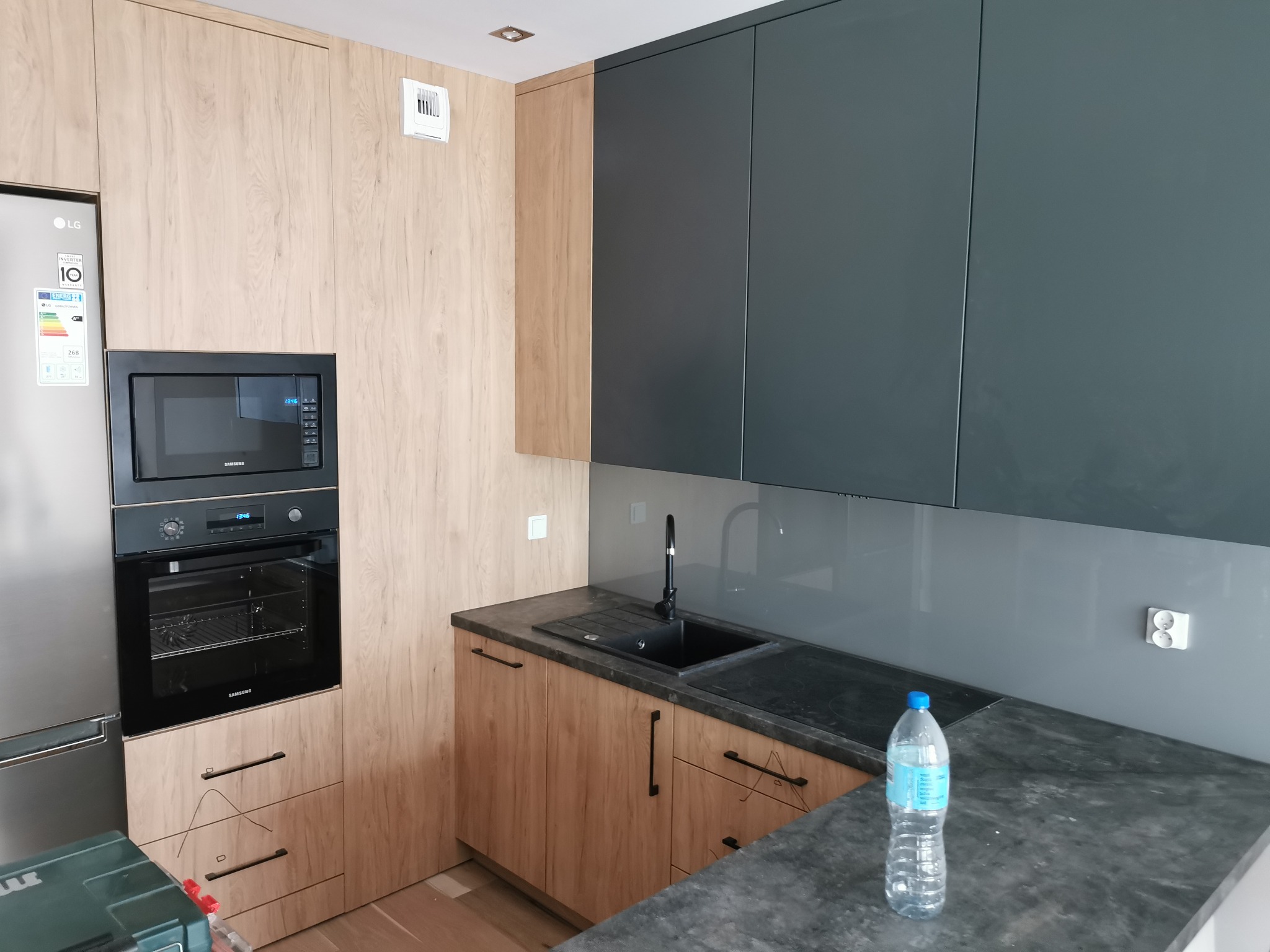 Nowoczesna kuchnia z drewnianymi frontami szafek dolnych i grafitowymi górnymi, czarny zlew i bateria, szary panel szklany nad blatem, wbudowany piekarnik i mikrofalówka Samsung, butelka wody...