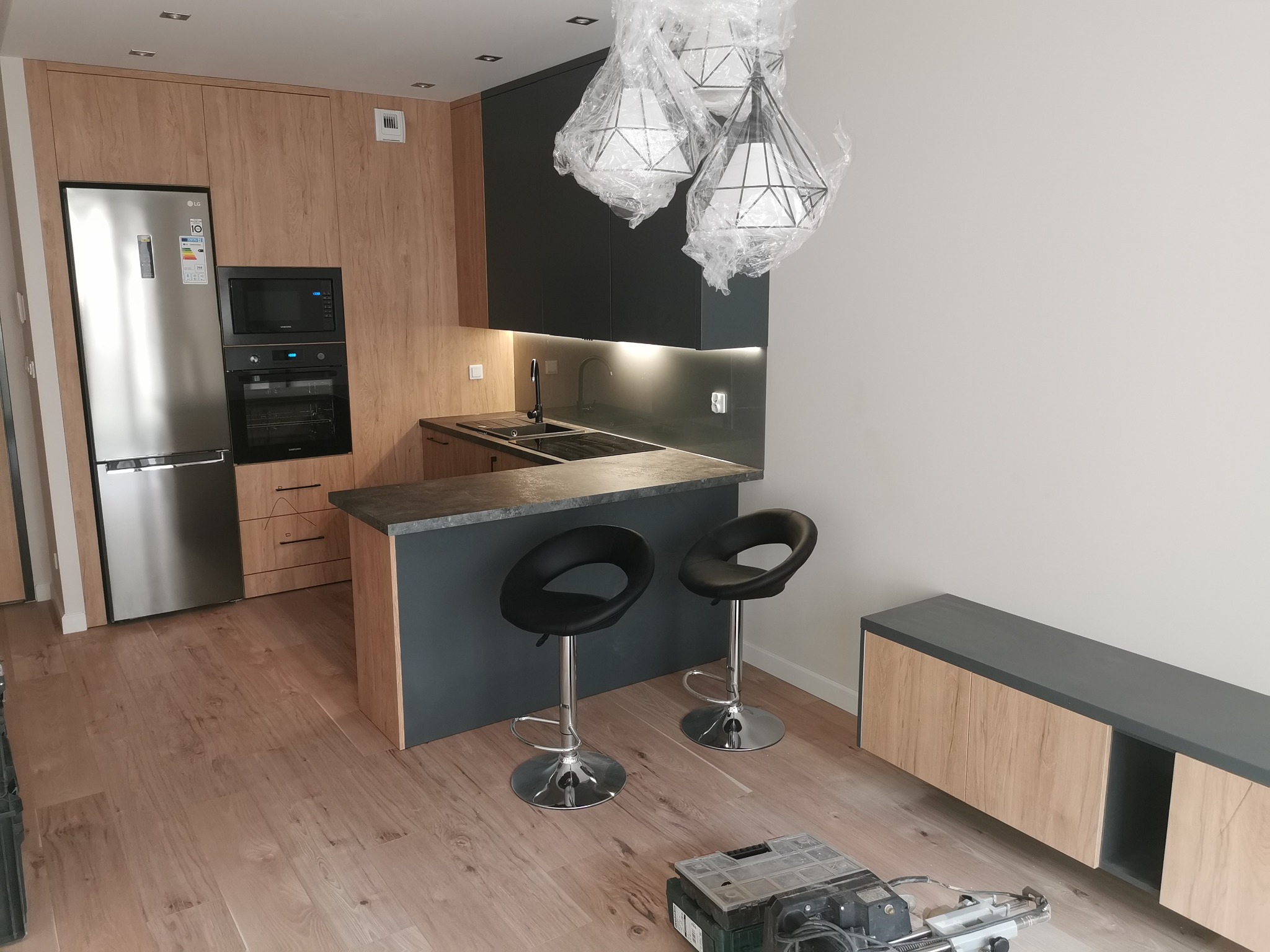 Nowoczesna kuchnia z drewnianymi frontami, czarnymi szafkami górnymi i barkiem, oświetlona trzema lampami w folii ochronnej, z dwoma czarnymi hokerami i widocznymi narzędziami na podłodze.
