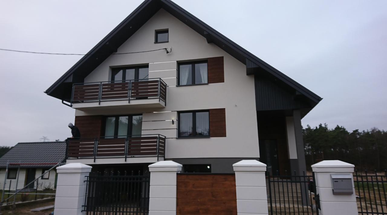 Dwukondygnacyjny dom z jasną elewacją, ciemnymi elementami wykończeniowymi, drewnianymi balustradami balkonów i ogrodzeniem z białymi słupkami.