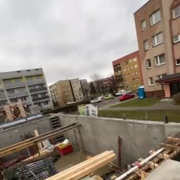 Usługi ogólnobudowlane Jakub Dyduch - Wysoki kąt ujęcia na plac budowy z widocznymi fundamentami, szalunkami i zbrojeniami na tle miejskiej zabudowy w pochmurny dzień. Widoczne materiały budowlane.