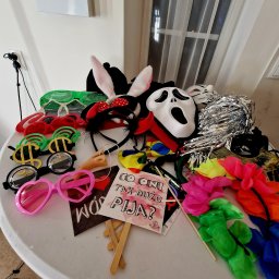 MZ Event Solutions - Biały stół z różnorodnymi rekwizytami: różowe okulary serca, zielone dolarowe, maska Scream, opaski z uszami królika i myszki, kolorowe naszyjniki kwiatowe i zabawne tabliczki.