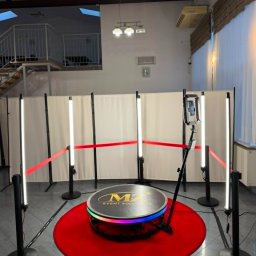 MZ Event Solutions - Gotowe stanowisko do fotobudki 360 z okrągłą platformą z logo, czerwonym dywanem, czterema pionowymi lampami LED i kamerą na statywie, otoczone czerwonymi linami.
