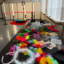 MZ Event Solutions - Stół z kolorowymi rekwizytami do zdjęć, takimi jak hawajskie naszyjniki, okulary i tabliczki z napisami, na tle fotobudki 360 z oświetleniem LED i czerwoną platformą.