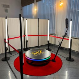 MZ Event Solutions - Platforma 360 z logo 'MZ EVENT SOLUTIONS' na okrągłym, czerwonym dywanie, otoczona pionowymi lampami LED i białymi parawanami, przygotowana do nagrywania wideo.
