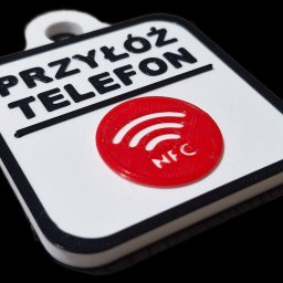Brylok - Biała, kwadratowa zawieszka z napisem 'PRZYŁÓŻ TELEFON' i czerwonym symbolem NFC, wykonana metodą addytywną. Widoczne warstwy materiału i czarne obramowanie.