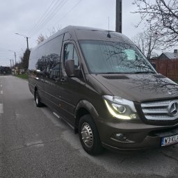 KLARIBUS DANIEL BRYL - Ciemnobrązowy Mercedes-Benz Sprinter z włączonymi światłami, zaparkowany na poboczu asfaltowej drogi. Widoczna tablica W3 KLARI, w tle wiejskie zabudowania i drzewa.