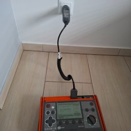 MW Elektro-System - Miernik Sonel MPI-530 podłączony do gniazdka elektrycznego na białej ścianie, na tle płytek podłogowych. Urządzenie w trakcie pomiaru impedancji pętli zwarcia.