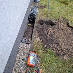 MW Elektro-System - Pomiar rezystancji uziomu przy budynku. Widoczny miernik, przewody pomiarowe, wykop w ziemi i wbity pręt uziemiający. Prace ziemne w trawie.