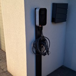 MW Elektro-System - Nowoczesna stacja ładowania EV, zamontowana na czarnym słupku przy ścianie budynku. Kabel zwinięty pod stacją. Obok skrzynka na listy w podobnym stylu.