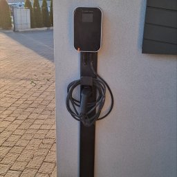 MW Elektro-System - Zewnętrzna stacja ładowania pojazdów elektrycznych zamontowana na ścianie budynku, z widocznym kablem i wtyczką. Obok fragment elewacji i kostki brukowej.