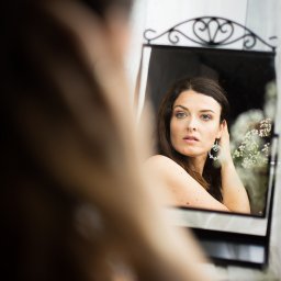AGACEBULAFOTO - Portret kobiety z kolczykami, odbijającej się w ozdobnym lustrze. W tle delikatne kwiaty i jasne światło. Ujęcie z bliska, z rozmytym pierwszym planem.