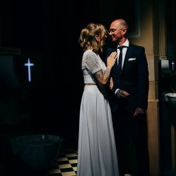 AGACEBULAFOTO - Para młoda w kościele, on w garniturze, ona w białej sukni i topie z koronki, oboje z tatuażami, patrzą na siebie w półmroku, na ścianie krzyż.