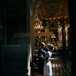 AGACEBULAFOTO - Para młoda w objęciach w kościele, widok przez krzyż na ścianie. Pan w garniturze, pani w białej sukni. W tle ołtarz i ławy.