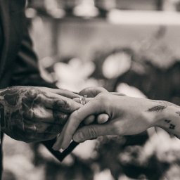 AGACEBULAFOTO - Czarno-białe zbliżenie dłoni z tatuażami, na które nakładany jest pierścionek zaręczynowy. Delikatne światło podkreśla detale biżuterii i wzory na skórze.