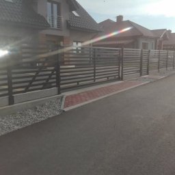 Marcin Pietrzyk - Nowoczesne, poziome ogrodzenie metalowe w kolorze grafitowym, z bramą przesuwną, na tle domu z balkonem i kostki brukowej. Widok z poziomu ulicy.