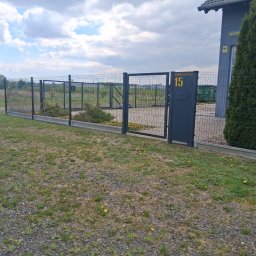 Marcin Pietrzyk - Nowoczesne ogrodzenie panelowe w kolorze grafitowym z furtką i numerem posesji '15' na tle zieleni i błękitnego nieba z chmurami. Widok z zewnątrz posesji.