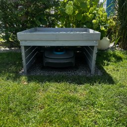 BS Home & Garden Solutions - Robot koszący Gardena w szaro-niebieskiej obudowie, zaparkowany pod drewnianą, jasnoszarą wiatą na żwirowym podłożu. W tle zielony trawnik, krzewy hortensji i wysokie trawy.