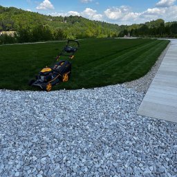 BS Home & Garden Solutions - Czarno-żółta kosiarka spalinowa stoi na świeżo skoszonym, pasiastym trawniku, obok ścieżki z jasnych otoczaków i betonowej płyty. W tle zielone wzgórza pod błękitnym niebem.