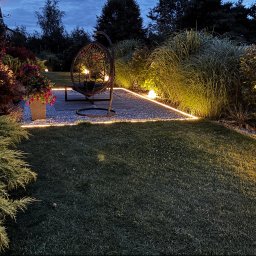 BS Home & Garden Solutions - Zadbany ogród o zmierzchu z podświetloną ścieżką żwirową, ozdobnymi trawami i krzewami. W centrum metalowy fotel wiszący typu kokon, otoczony ciepłym światłem.