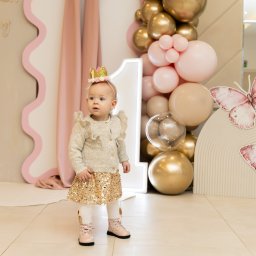 Karina Zioła Fotografia - Roczne dziecko w koronie pozuje na tle dekoracji urodzinowych z balonami, motylami i cyfrą '1' w jasnych, pastelowych kolorach. Dziewczynka ubrana w sweterek i spódniczkę.